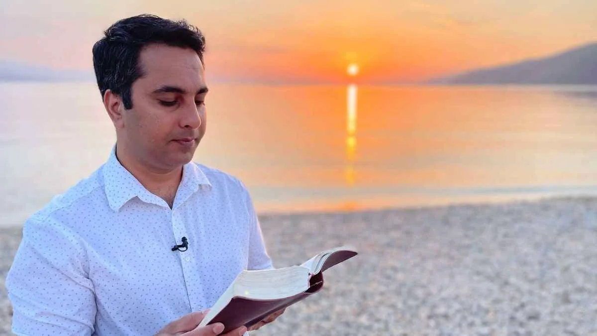 Man reading an open book beside a lake at sunset.