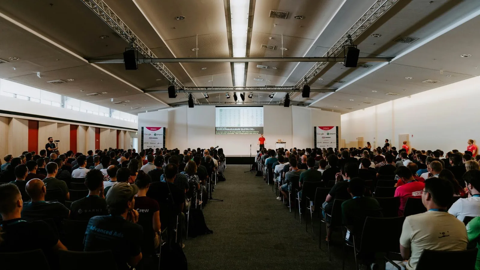 Audience seated in a conference hall facing a speaker and projection screen.