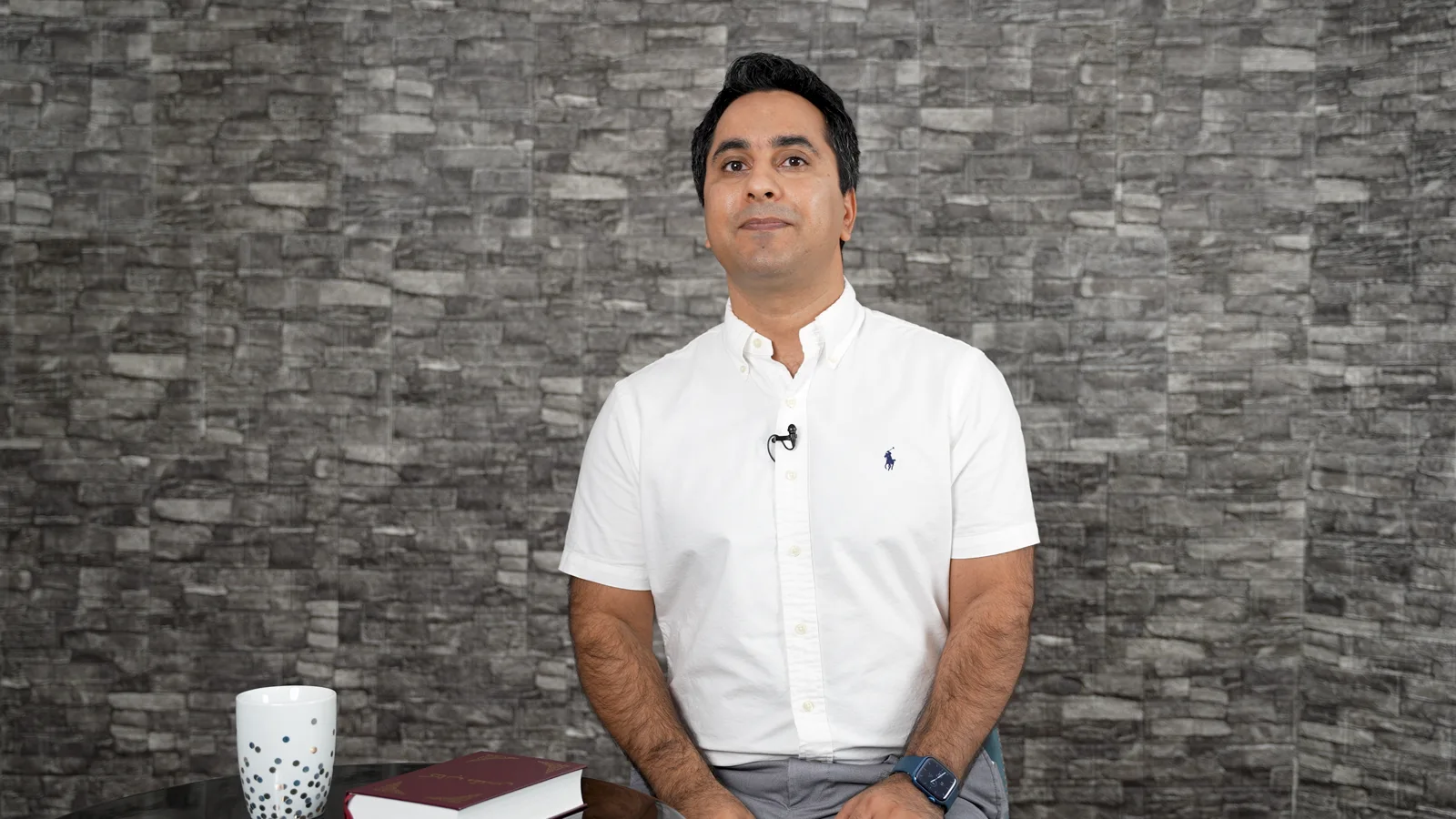 Man in a white polo shirt seated beside books and a mug against a stone-patterned wall.