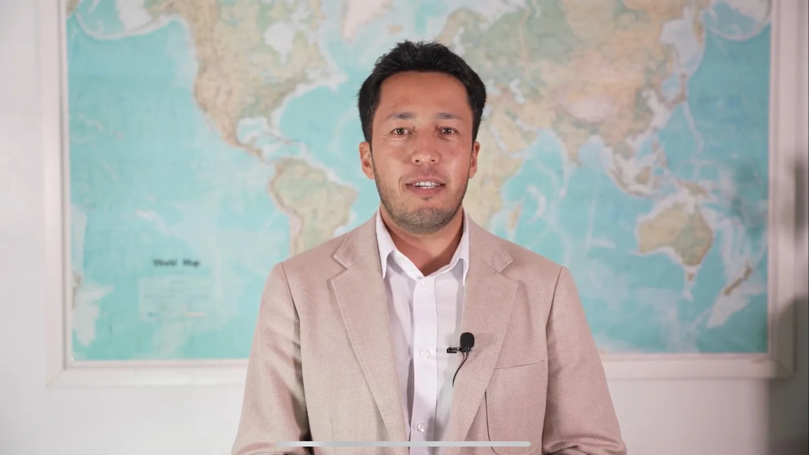 Man in a beige jacket seated in front of a world map.